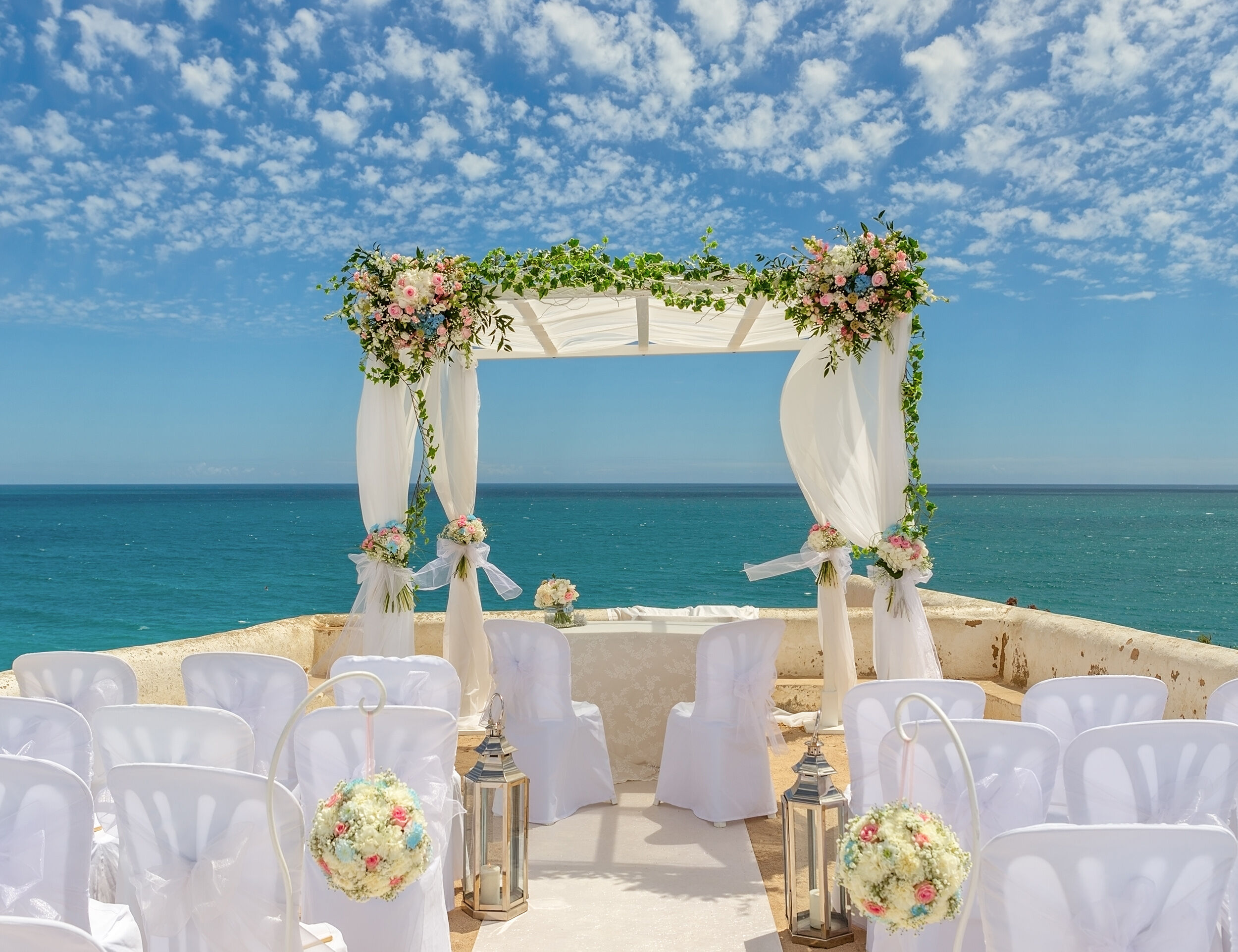 Elegant seaside wedding setup with white draped mandap, pastel floral decorations, and white chairs arranged under a bright blue sky with scattered clouds.